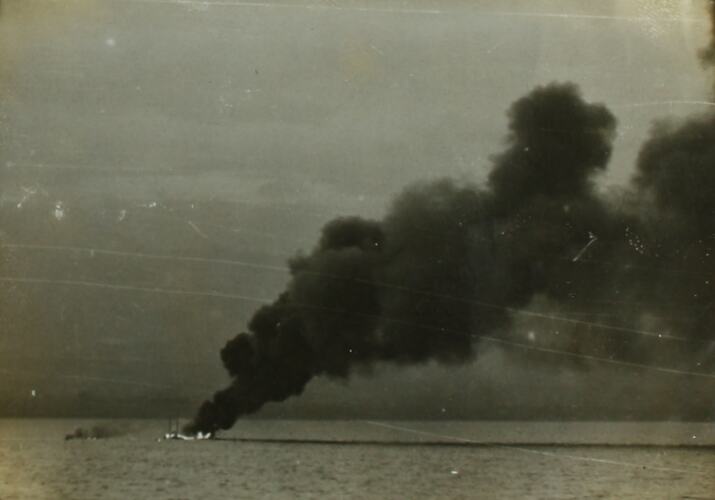 Black plume of smoke rising from ship on fire in ocean.