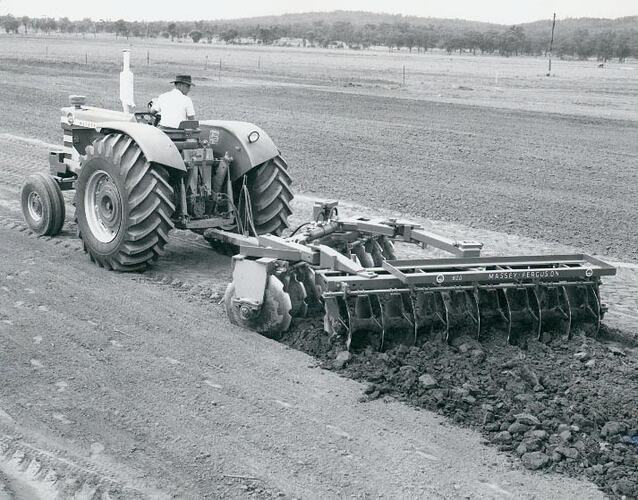Rear left view of a man driving a tractor coupled to a harrow in a field.