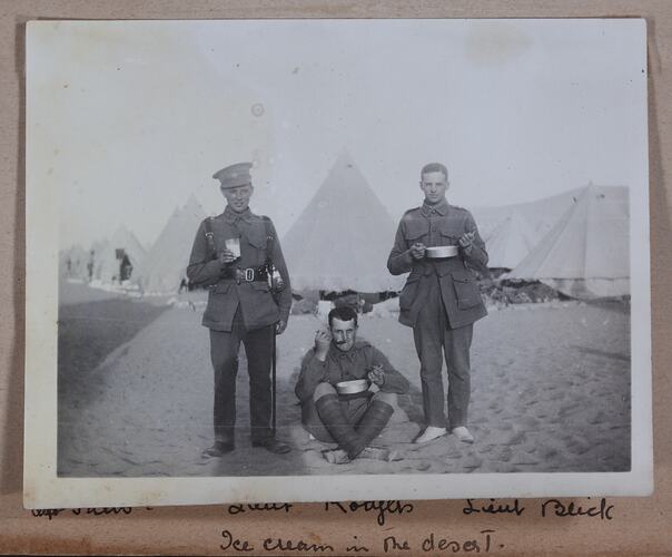 Two servicemen standing and one sitting in front of tents on sandy ground.