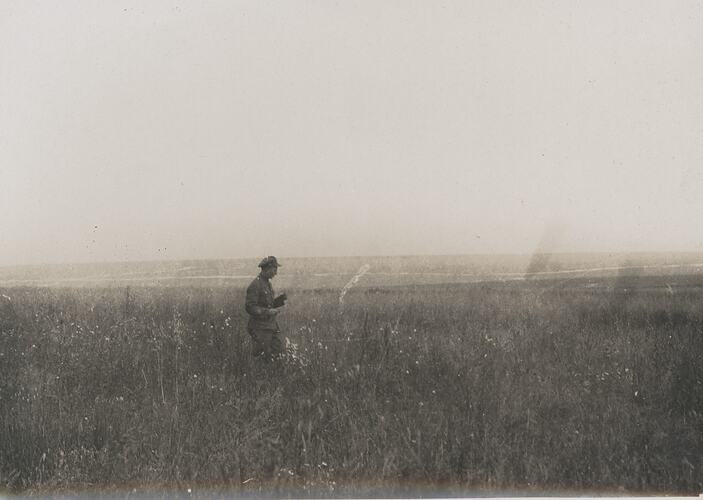 Man in military attire stands in an open, flat field with grass up to his knees.