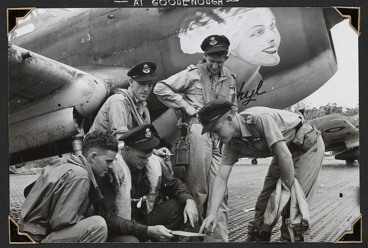 Group of serviceman next to aircraft looking at board, painting of woman's head on side of aircraft.