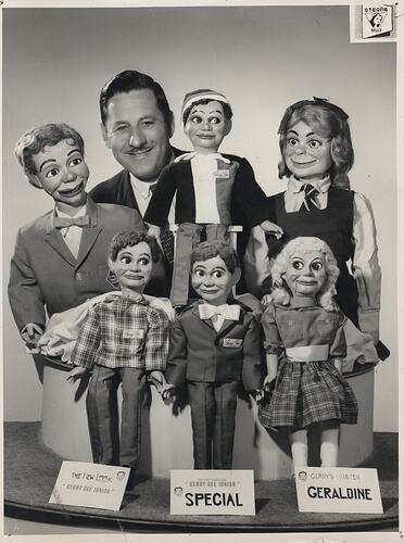 Black and white photograph of a person standing amongst ventriloquist dolls.