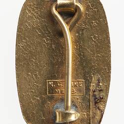 Back of rectangular shaped metal badge with rounded corners. Has pin and hook and maker's name.