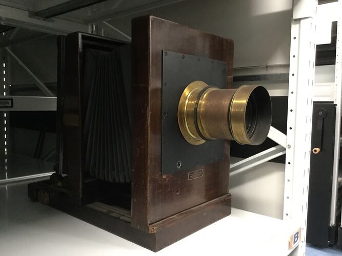 Box-like wooden view camera with large brass lens on a fixed front standard.