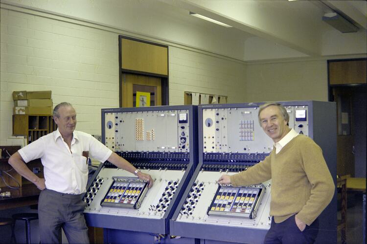 Two men pose in front of two large computers.