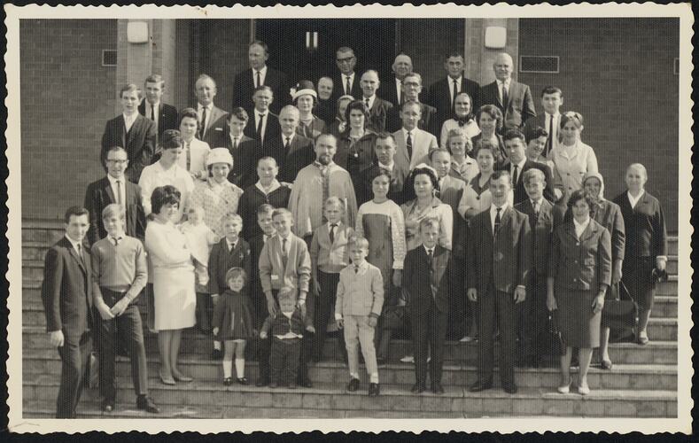 Group portrait of approximately 50 people posing on steps. Priest stands near centre.