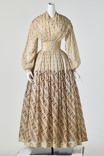 Cream muslin dress, high neck, long sleeves, banded waist, full length patterned gathered skirt.