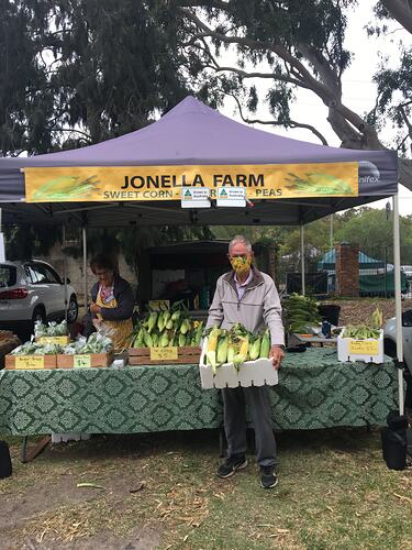 Man wearing a sweet corn mask standing in front of a corn stall holding a box of corn.