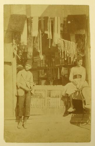 Shopfront with two young men, one standing, the other sitting on a ledge.