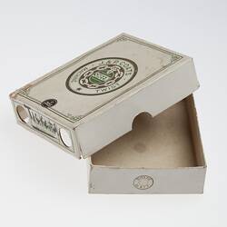 White card box with partially removed lid. Central circular logo with text in gold and green ink.