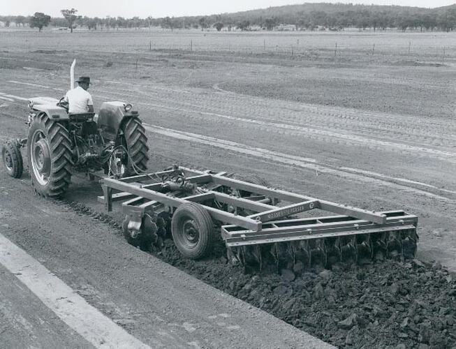 Rear left view of a man driving a tractor coupled to a harrow in a field.
