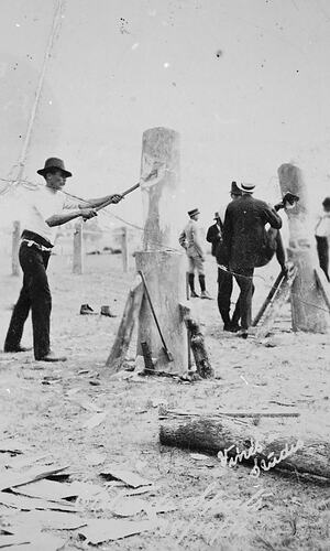 Wood chopper striking a tree trunk. Men stand around a second trunk behind him at right. More trunks behind.