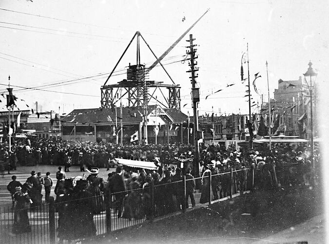 Flinders Street Railway Station under contruction. Large crane on platform in background. Crowds line street, bunting on poles.
