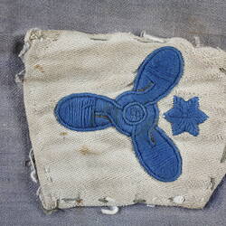 Worn, stained, off-white rectangular badge with central embroidered blue propeller above blue star.