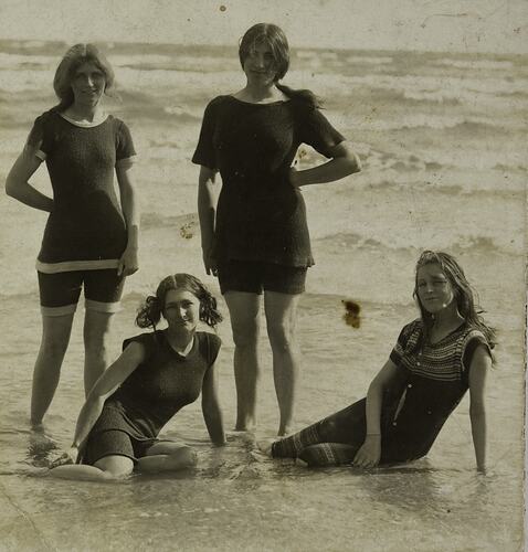 Four young women pose on beach shoreline wearing older style swimwear. Short sleeve tops and shorts.