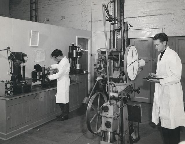 2 men in white coats, standing at testing equipment.