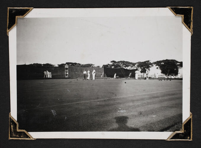 Men playing lawn bowls, with tarp fencing and trees in background.