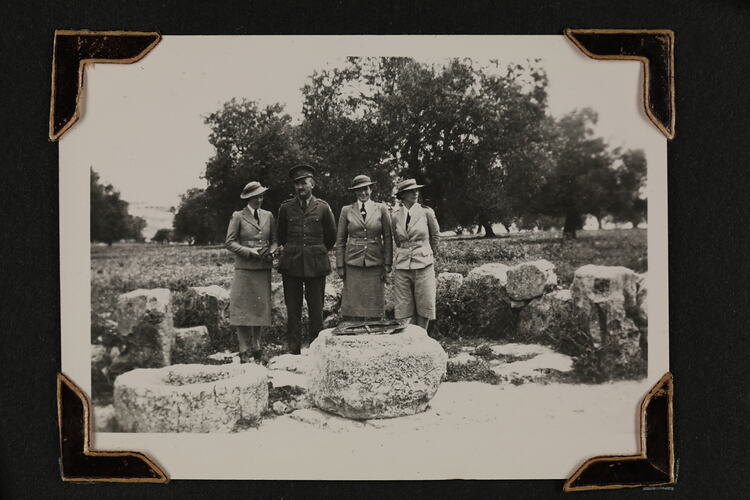 Three woman and one man in uniform standing by a stone well, with stones in a row  behind.