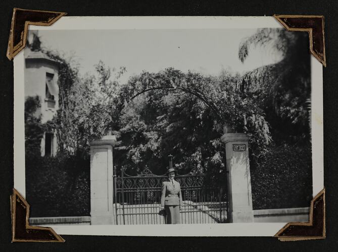 Woman in uniform standing in front of gate, with garden and house behind.