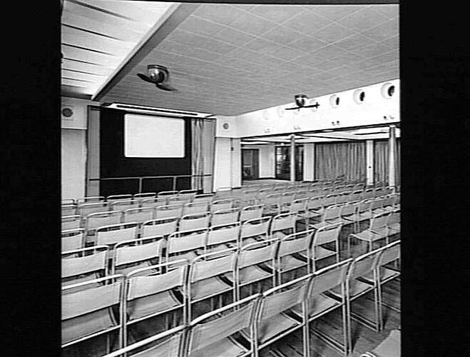 Ship interior. Room with metal chairs set up in rows to create cinema seating. Projector screen on back wall.