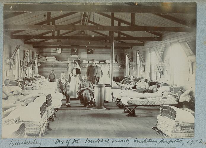 Hospital ward with people in beds on either side and group on people posed in centre.