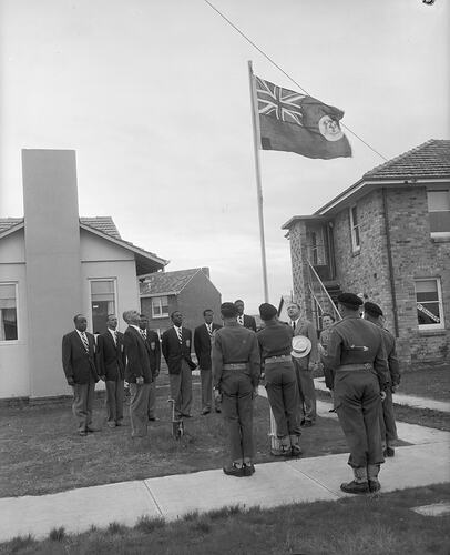 Sportsmen wearing blazers standing to attention around a raised Colonial Flag of Jamaica. Residential buildings in background.
