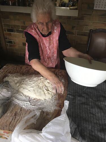 Woman making bread dough on wooden bench. Holds white bowl in her left hand.