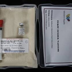 Labelled syringe and vial in lined plastic box. Card in lid has printed text. Black background.