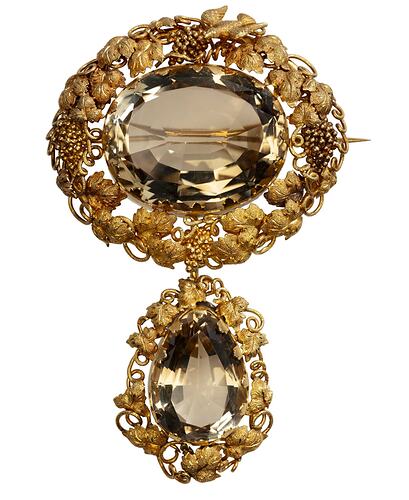 Two part brooch. Top, ornate gold frame with transparent brown cut stone. Below hangs smaller teardrop version