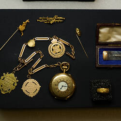 Gold items including a pin, medals, fob watch and ring on black display board.