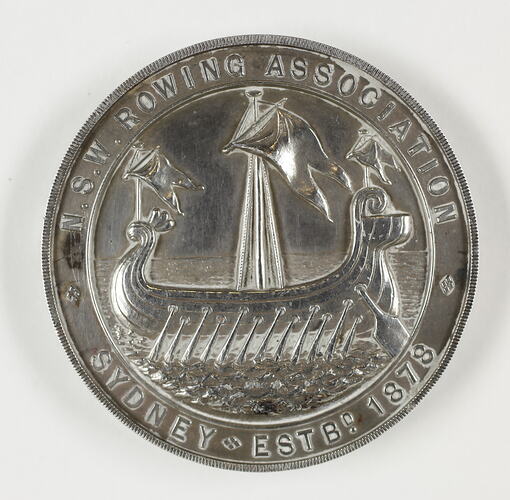 Round medal with rowing ship framed by border and text.