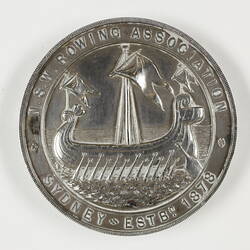 Round medal with rowing ship framed by border and text.