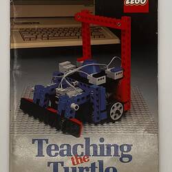 Front cover with LEGO build. Blue and red, grey base. Wheeled, grey cable. Text above, below. Computer behind.