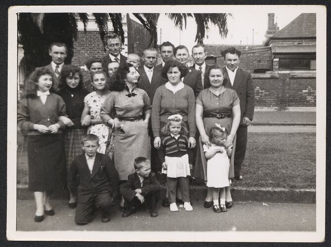 Group of approx 20 men, women and children pose in front of brick wall beneath a palm tree.