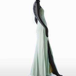 Full length pale green silk evening slip dress. Sleeveless. Fastens at back with long green silk covered rope ties. Flared/fishtail skirt with long slit at right. Right profile.