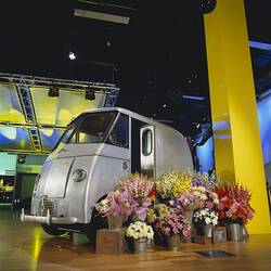 Small silver van on display indoors with flowers in buckets in front of it.