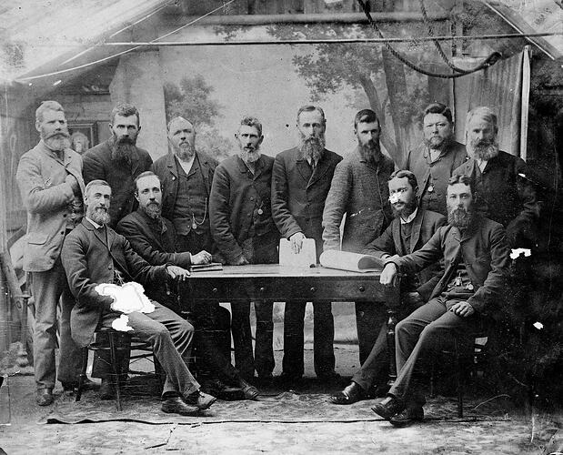 Group portrait of 12 men. Some sit, others stand around a table. Decorative backdrop screen behind them.