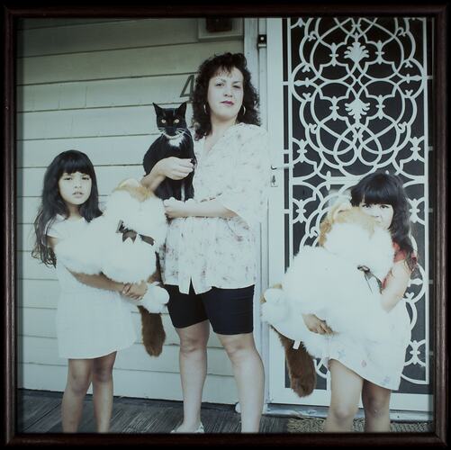 Woman standing on veranda holding a cat. A young girl stands either side of her holding soft toys.