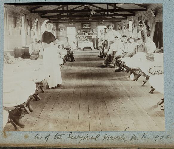 Hospital ward with people in beds on either side and others posed standing or sitting next to beds.