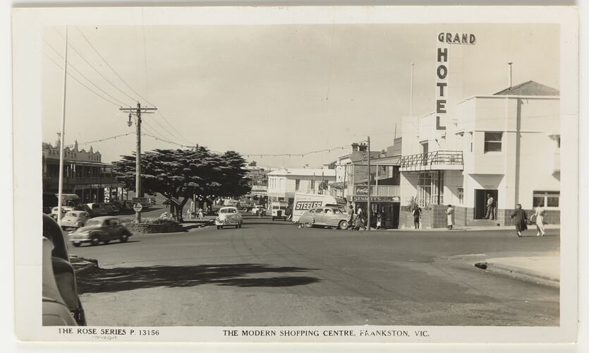 Streetscape of a cars on a road in a shopping strip. Trees at left and hotel at right.
