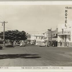Streetscape of a cars on a road in a shopping strip. Trees at left and hotel at right.