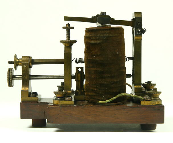 Brass apparatus with battery in centre on wooden base, side view.