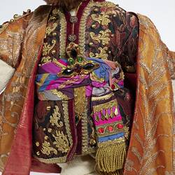 Detail of light brown plush bear wearing ornate burgundy and gold tunic, purple blue belt and orange gold coat