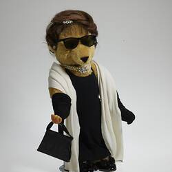Light brown bear wears black hat, dress, full-length gloves, shoes and white scarf.