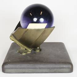 Purple glass sphere sitting on metal frame with square base, side view.