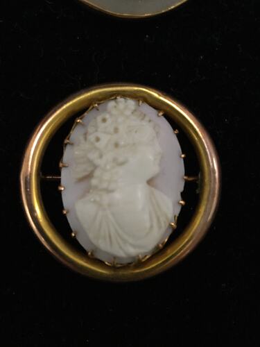 Round cameo brooch, white female bust facing right, black background. Gold border.