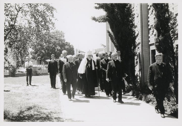 Cardinal, bishop and other men outside a brick building, walking along side it. Image of them walking taken from the front.