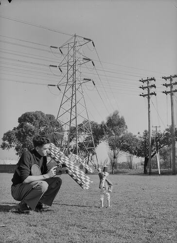 Man squatting on grass with puppet and model aircraft near overhead transmission lines.