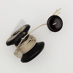 Black wooden cotton reel with two nails in top. White string tied around the middle. The string has a black button on the end.