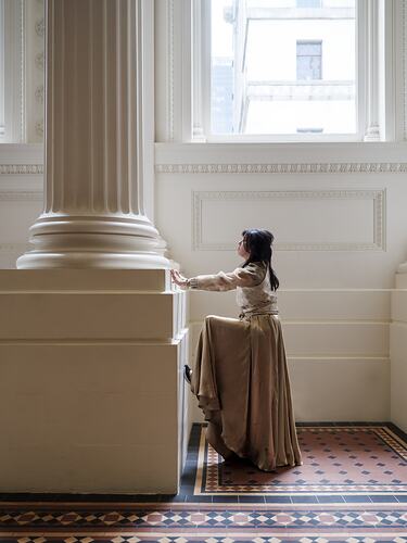 Profile of woman in long pale shirt and skirt pushing against column base.